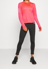 Maglietta sportiva a maniche lunghe di colore rosa con inserti in rete e tre strisce bianche sul braccio sinistro, abbinata a leggins neri con polsini con zip.