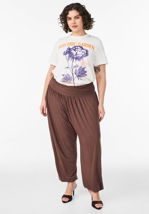 Kobieta z kręconymi włosami, ubrana w białą koszulkę z kwiatowym nadrukiem "Electric Garden", luźne brązowe spodnie i czarne sandały na obcasie z odkrytymi palcami, stojąca na białym tle.