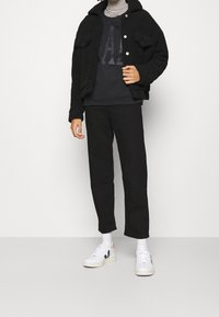 Chaqueta negra de felpa con cierre de botones y dos bolsillos en el pecho, sobre un sudadera oscura con un gráfico grande. Combinada con pantalones negros y zapatillas blancas.