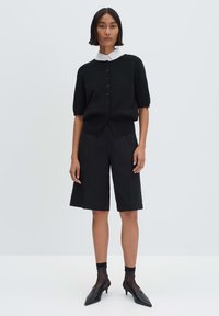 Black short-sleeve cardigan with buttons, white collared shirt underneath, paired with black tailored shorts and pointed black heels.