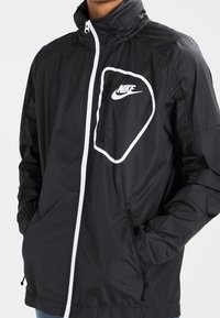 Svart vindjacka med en vit Nike-logotyp på bröstfickan, framdragkedja, justerbar krage och sidofickor.
