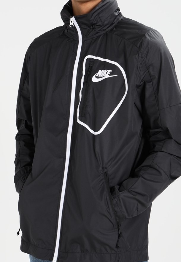 Svart vindjacka med en vit Nike-logotyp på bröstfickan, framdragkedja, justerbar krage och sidofickor.