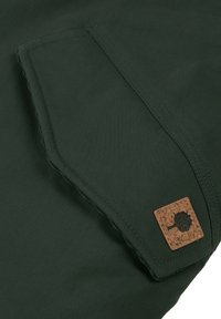 Groene stof met een geweven textuur; heeft een opgestikte zak en een vierkante kurklogo-accent. Schoon, recht gestikte naden langs de randen.