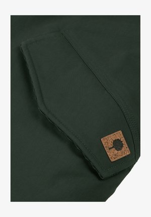Tessuto verde con una trama intrecciata; presenta una tasca a toppa e un accento in sughero a forma quadrata. Cuciture pulite e dritte lungo i bordi.