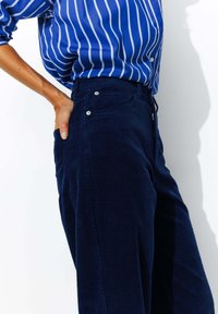 Pantalons en velours côtelé bleu marine avec une surface côtelée texturée et des poches latérales, associées à une chemise rayée bleue à coupe décontractée et fermeture à boutons.