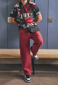 Czarna koszulka polo z czerwonym napisem "Forza Milan", zestawiona z luźnymi czerwonymi spodniami i czarnymi trampkami z białymi akcentami. W tle szafki.