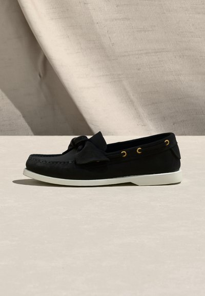 Μαύρο μοκασίνι slip-on με λευκή σόλα, πλευρικές οπές και μεγάλο φιόγκο στην κορυφή, εμφανίζεται σε ουδέτερη επιφάνεια με φόντο υφάσματος.