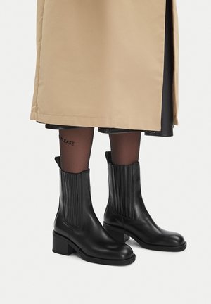 Czarne skórzane botki na słupku, noszone z czarnymi, przezroczystymi rajstopami i beżowym płaszczem trenczowym na czarnej spódnicy.