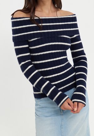 Femme portant un pull à rayures bleu marine et blanches, manches longues, épaules dénudées, et une jupe en denim bleu clair, les mains jointes devant elle.