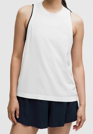 Kobieta w białym sportowym topie bez rękawów, na czarnym sportowym biustonoszu i czarnych sportowych szortach na jednolitym tle.