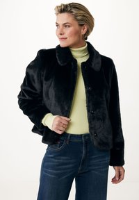 Svart fuskpälsjacka med krage, tryckknappar och figursydd design. Bärs över en ljusgrön polotröja och blå jeans.