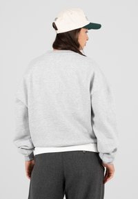 Fiatal nő oldalra néz, világosszürke pulóvert, sötétszürke nadrágot és fehér-zöld baseball sapkát visel.