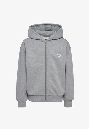 Barn står med front mod kamera iført en grå hættetrøje med lynlås, matchende bukser med vide ben, hvide sneakers og en mørkegrøn hue. Hænderne i lommerne.