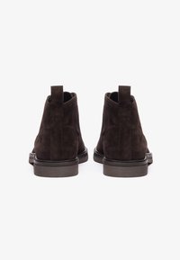 Botines de ante marrón con una textura suave, puntera redonda y tiradores en la parte trasera. Suela de goma negra con un leve tacón.