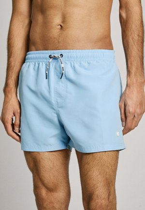 Shorts de bain bleu clair avec taille élastique, cordon de serrage et un petit logo en bas à gauche. Texture lisse et longueur au-dessus du genou.
