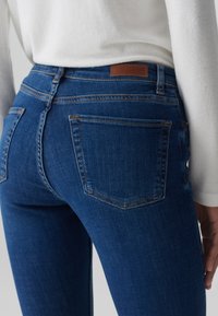 Jeans skinny blu in denim, con vita media, due tasche posteriori e cuciture visibili di un colore contrastante. Etichetta in pelle marrone in vita.