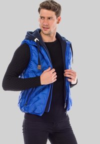 Blauer Puffer-Westen mit Kapuze, Reißverschluss vorne und kontrastierendem dunklem Innenfutter. Verfügt über gerippte Strickärmel und eine glatte Textur.