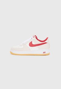 Teniske Nike Air Force 1 iz bež semiša z rdečimi akcenti, opremljene z belimi vezalkami, perforiranim prstom in gumijastim podplatom.
