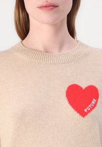 Beżowy sweter wykonany na drutach z okrągłym dekoltem, z dużym czerwonym sercem wszytym na klatce piersiowej oraz słowem "FUTURE" w białym kolorze.