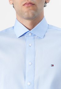 Lys blå skjorte laget av glatt stoff, med klassisk krage, hvite knapper, og en liten brodert logo på brystet.