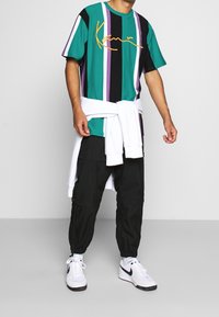 T-shirt z krótkim rękawem w paski w kolorze turkusowym, czarnym i purpurowym, z żółtym tekstem napisanym kursywą. Połączony z czarnymi spodnimi spodniami i białymi sneakersami.