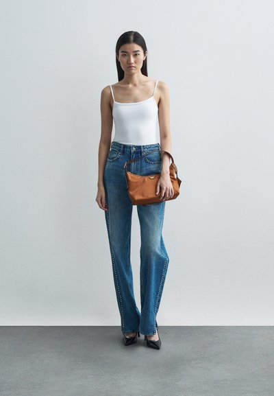 Kvinna i vit linne och blå jeans med nitar på sidorna, håller en brun handväska, stående på grått golv mot en vit vägg.