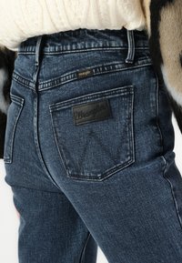 Tamno plavi traper hlače s crnom kožnom oznakom na kojoj je ispisano "Wrangler." Karakteristike uključuju visoki struk i standardne stražnje džepove.