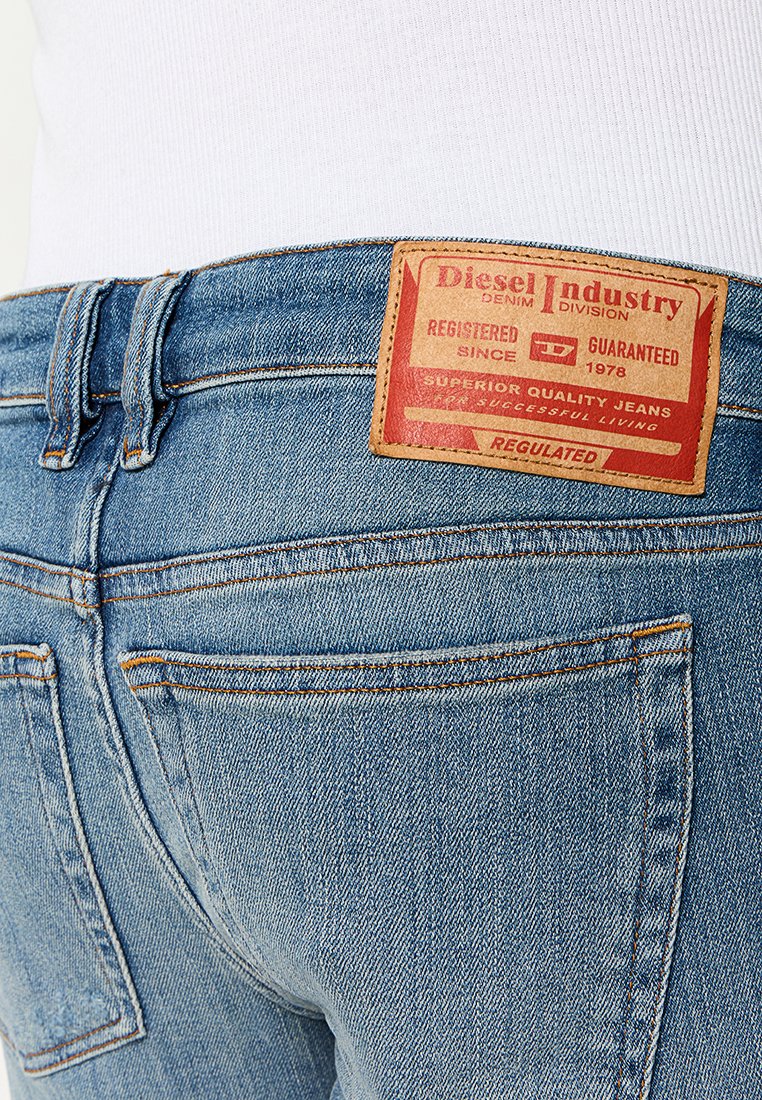 Jean en denim bleu clair avec une coupe droite, présentant une pièce en cuir marron sur la ceinture arrière. Les coutures sont d'une couleur orange contrastante.