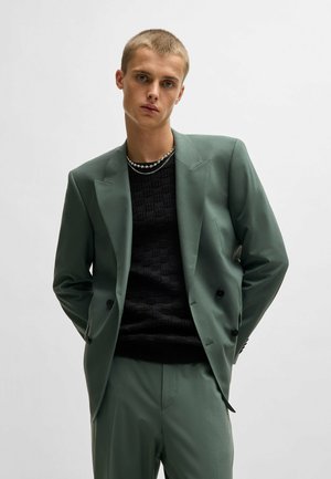 Dubbelknäppt grön kavaj i slätt tyg, kombinerad med en texturerad svart tröja. Modellen poserar med händerna i fickorna, neutral bakgrund.