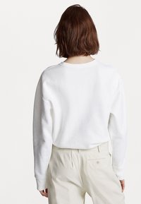 Person med kort brunt hår som bär en enkel vit långärmad tröja och beige högmidjade byxor, visad bakifrån.