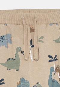 Pantaloni beige per bambini con vita elastica e cordoncino, caratterizzati da motivi giocosi di dinosauri e palme in tonalità di blu, verde e marrone.