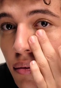 Primer plano de un rostro que destaca una piel suave, ojos marrón oscuro y un color de labios natural. Una mano está tocando suavemente la piel cerca del ojo.