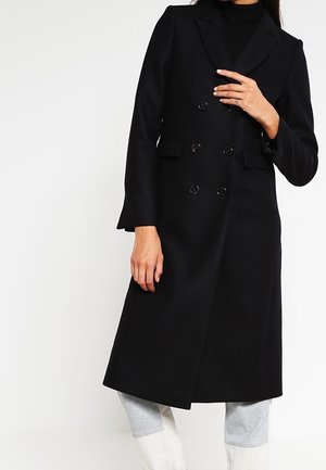 Manteau noir à double boutonnage avec revers crantés, tissu en laine texturé, poches latérales et quatre boutons ronds noirs sur le devant, longueur aux genoux.