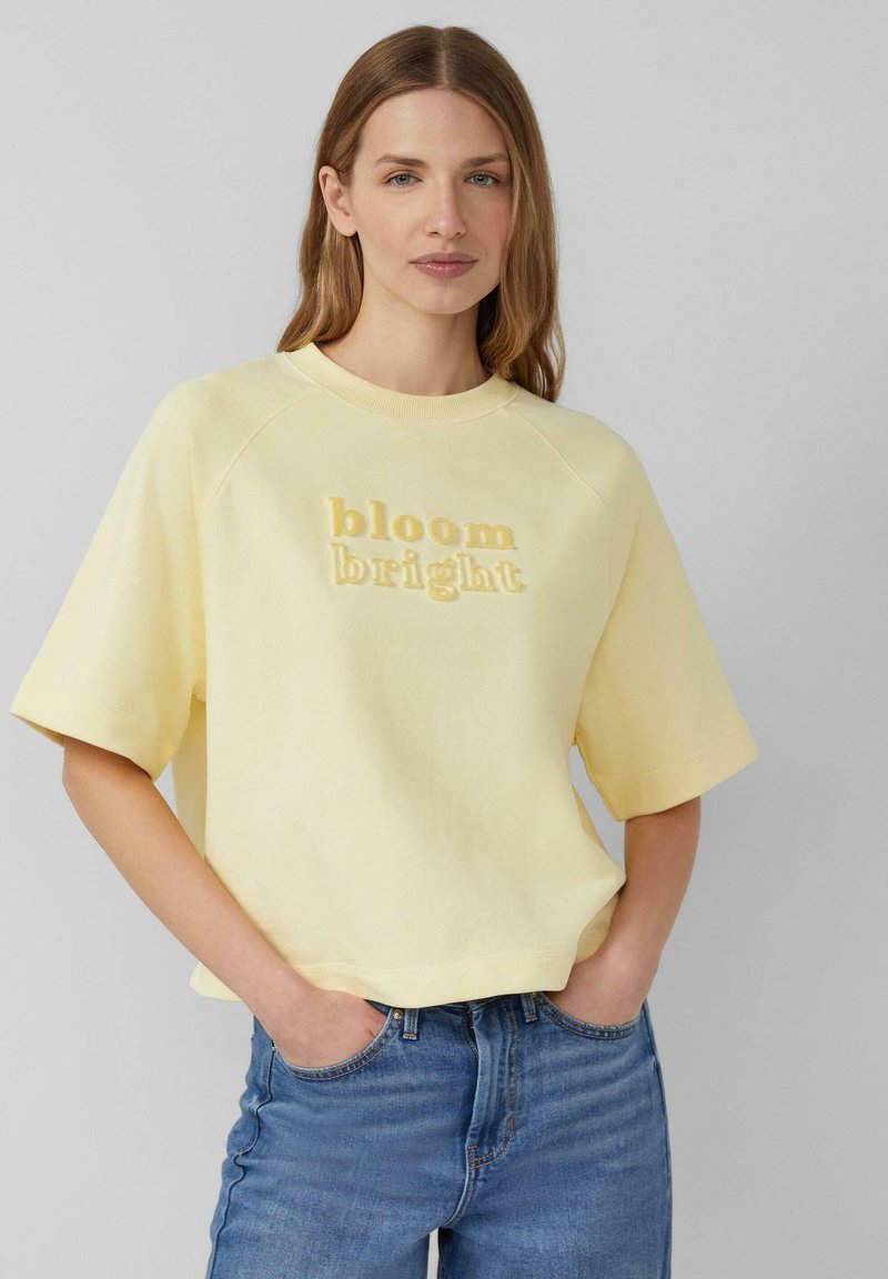 Kobieta o prostych, jasnobrązowych włosach nosi bladoróżowy krótki rękaw z napisem „bloom bright” oraz niebieskie dżinsy, ręce w kieszeniach.