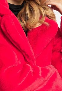Cappotto in finta pelliccia rossa con un ampio colletto e una consistenza morbida e spessa. Presenta rever oversize e dettagli di cucitura visibili lungo i bordi.