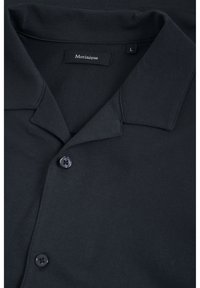 Svart pikétröja med kort krage, två svarta knappar och en etikett med texten "Martinique." Tyget verkar slätt med en lätt glans. Storlek L.