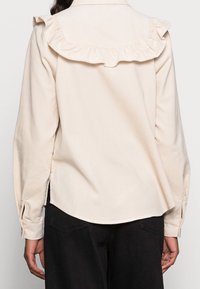 Hellbeige Bluse mit Rüschenausschnitt, langen Ärmeln, seitlichen Schlitzen und Knopfmanschetten. Glatte Stoffqualität mit lockerer Passform und abgerundetem Saum.