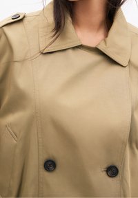 Olivengrøn trenchcoat med dobbeltradet design, store sorte knapper og bred krave med strukturerede skuldre.