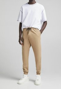 Weißes T-Shirt mit kurzen Ärmeln und grauen Nähten, kombiniert mit beigen Joggerhosen mit elastischem Bund und Bündchen. Weiße Sneakers runden das Outfit ab.