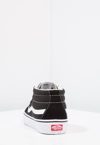 Schwarze High-Top-Sneaker aus Canvas mit weißem Logoakzent, weißen Nähten und einem roten Logo auf der Ferse. Gummisohle für besseren Halt.