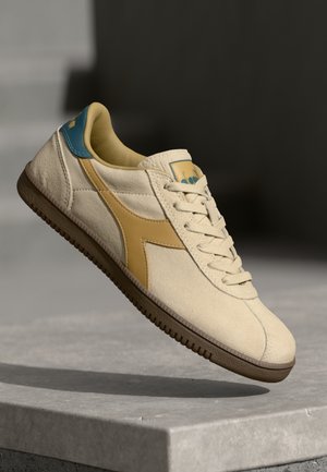 Beige tenisky s texturovaným povrchem, s žlutými a modrými akcenty, polstrovaným límcem a hnědou gumovou podrážkou.