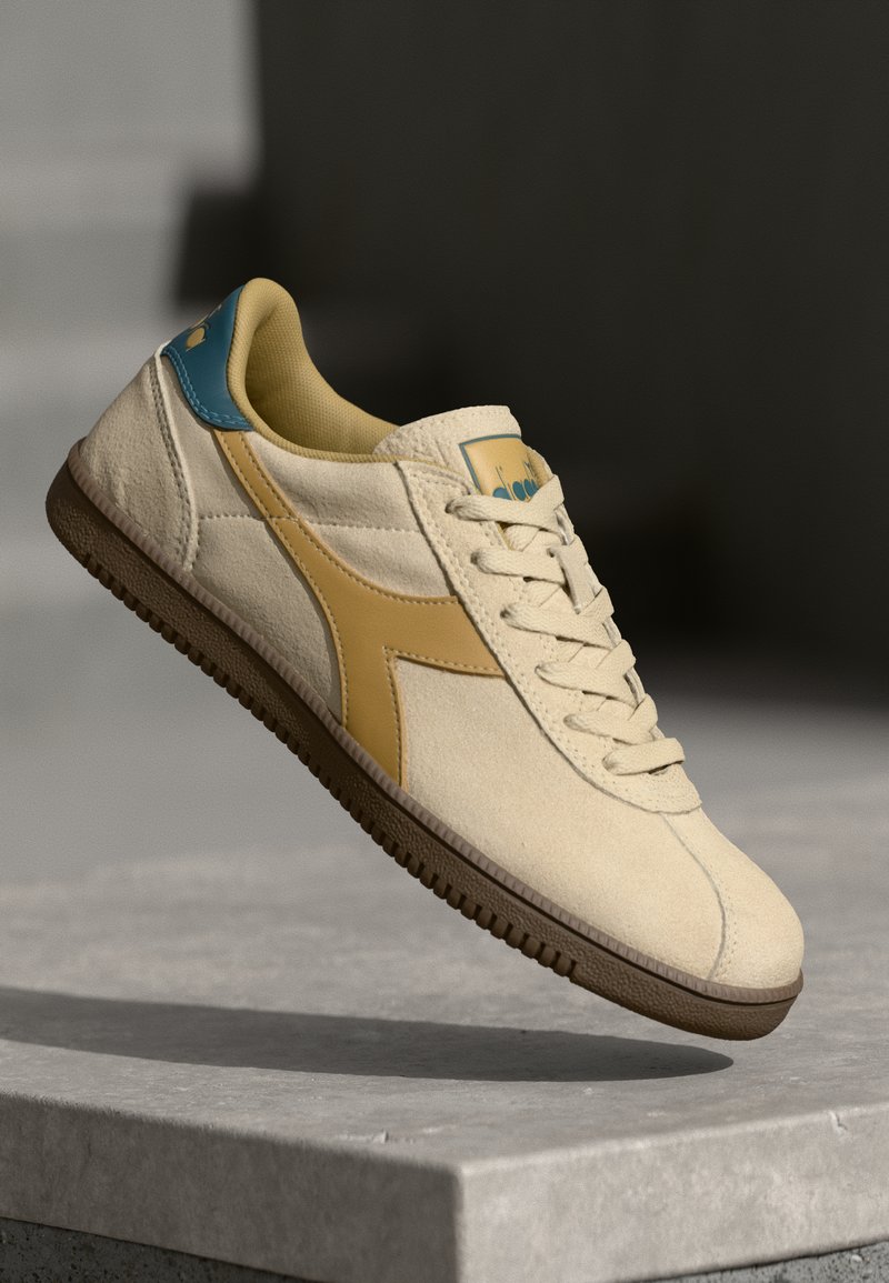 Sapatilha bege com uma superfície texturizada, apresentando detalhes em amarelo e azul, um colarinho acolchoado e uma sola de borracha castanha.