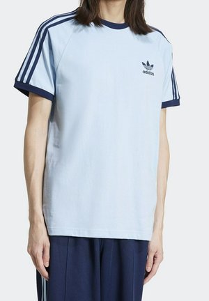 Hellblaues Baumwoll-T-Shirt mit marineblauen Akzenten, mit einem Rundhalsausschnitt und drei marineblauen Streifen auf jeder Schulter, sowie einem kleinen schwarzen Adidas-Logo auf der Brust.