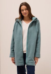 Vaaleansininen fleece-takki, jossa on vetoketjukiinnitys, huppu ja sivutaskut. Pehmeä rakenne ja rento ylipituinen leikkaus. Eri väriset reunukset korostavat muotoilua.