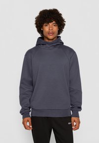 Felpa con cappuccio grigio scuro, colletto alto, polsini a coste, maniche raglan e una consistenza morbida. Indossata con pantaloni sportivi neri.