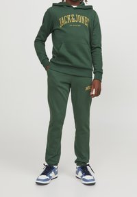 Grön hoodie och joggers-set från JACK & JONES. Har en gul textlogotyp, ribbade ärmmuddar och avslappnad passform. Bärs med blå och vita sneakers.