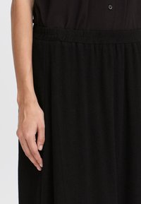 Jupe noire avec une taille élastique, présentant une texture lisse et un design fluide. Une main est posée sur le tissu.
