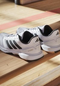 Chaussures de sport blanches avec des accents noirs, texture en maille et semelles rembourrées. Présente la marque "LIGRA" et trois bandes sur les côtés.