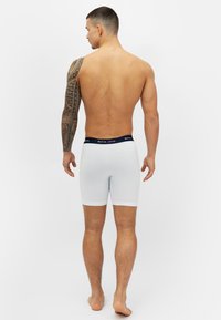 Valkoiset puuvillabokserit, joissa on tummansininen vyötärönauha, sileä pinta ja joustava istuvuus, nähtynä takaa henkilön tatuoidusta kädestä.