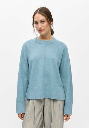 Jonge vrouw met lichtbruin haar in een losse knot, draagt een lichtblauwe gebreide trui en lichtgrijze wijde broek, staand tegen een witte achtergrond.